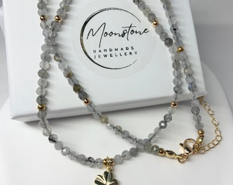 Peut inclure: Un collier délicat en argent avec un pendentif trèfle à quatre feuilles. Le collier est composé de petites perles facettées et d'accents dorés. Le collier est présenté sur une boîte blanche avec le texte "Moonstone Handmade Jewellery".