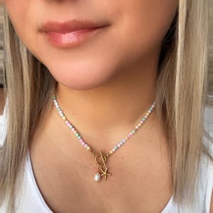 May include: A pastel beaded necklace with a gold clasp and charm. The charm includes a starfish and a pearl pendant. The necklace is worn on a person wearing a white tank top.