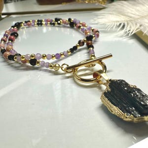 May include: A layered necklace with a gold clasp and a black stone pendant. The necklace features small, colorful beads in shades of pink, purple, black, and gold. The pendant is a rough-cut, dark stone with gold edging.