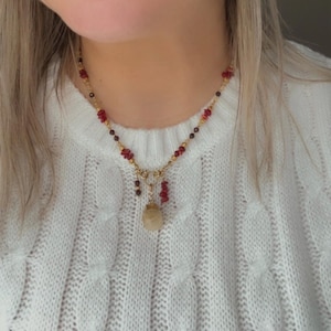 Op de afbeelding: Een goudkleurige ketting met rode en bruine kralen en een hanger. De ketting wordt gedragen over een witte kabelgebreide trui. De hanger is een lichtgekleurde, druppelvormige steen.