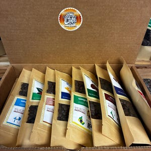 May include: A cardboard box filled with several sealed bags of coffee beans. Each bag has a label with text and a colorful design. The bags are arranged neatly inside the box. A circular sticker with text is on the box lid.