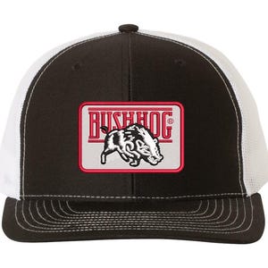 May include: Black and white trucker hat with a white mesh back. The front of the hat features a red and white embroidered patch with a wild boar and the text "BUSHHOG".
