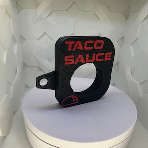 Puede incluir: Un soporte de calibre de plástico negro con una inscripción roja "TACO SAUCE". El soporte tiene una abertura redonda y una pequeña manija lateral.