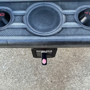 May include: A black plastic bumper with two round black emblems that say "T R O" in red letters. The bumper has a black hitch receiver with a pink release lever and a white sticker that says "WATCH YOUR SHIN".