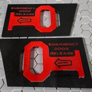 May include: Two black and red emergency door release signs. The signs have the words "EMERGENCY DOOR RELEASE" in red, along with a red arrow and a red key-shaped design. The signs are on a gray and white tiled surface.