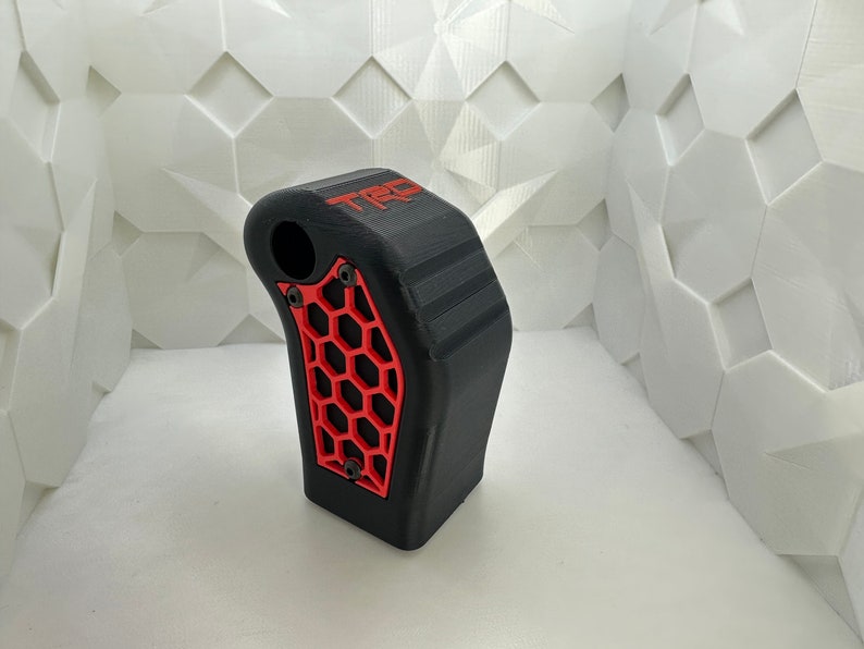 May include: Black and red 3D printed gear shift knob with a honeycomb pattern and the word "TRD" printed on the side.