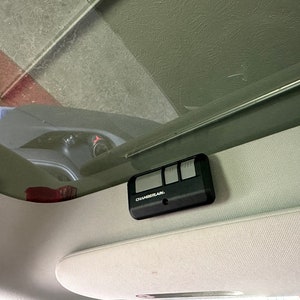 May include: A black garage door opener with three buttons and the text "CHAMBERLAIN" is mounted on the ceiling of a car.