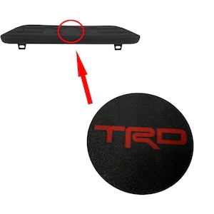 May include: Black plastic car emblem with a red "TRD" logo.