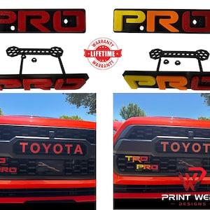 May include: Red and yellow "PRO" emblems and a black bracket with the text "WARRANTY LIFETIME WARRANTY". The emblems are shown installed on a red Toyota truck grill. The text "TOYOTA" is also visible.