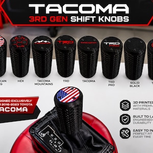 May include: A collection of eight black 3D-printed shift knobs for 2016-2023 Toyota Tacoma vehicles. The knobs feature textured sides and various top emblems including the American flag, hex patterns, Tacoma mountains, TRD, and Toyota logos.