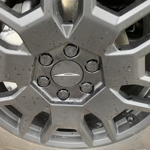 May include: Close-up of a black car wheel with a geometric design. The wheel has a central hubcap with a white logo and is surrounded by six lug nuts, each with a small logo. The tire is visible at the bottom.