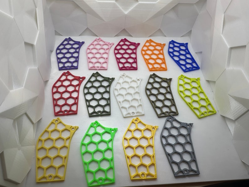 May include: A collection of 15 plastic honeycomb-shaped pieces in various colors, including red, purple, pink, orange, yellow, green, white, and gray. The pieces are arranged in rows on a white surface.