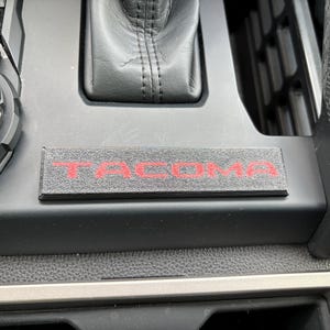 以下が含まれることがあります： 赤色の「TACOMA」の文字が刻まれた、長方形のテクスチャ加工された表面を持つ黒い車の内装コンソール。コンソールには、ギアシフト、コントロールダイヤル、模様入りのトリムが備わっています。