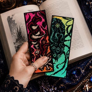 May include: Two rectangular cross-stitch bookmarks featuring intricate needlework designs. One bookmark displays a vibrant pattern of pink, red, and orange threads on a black background, while the other showcases a teal and green creature motif.