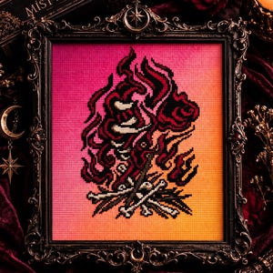 May include: A cross-stitch embroidery featuring a stylized bonfire with flames in shades of red and black, set against a vibrant pink to orange gradient background, all encased in an ornate, dark, metallic-toned rectangular frame.