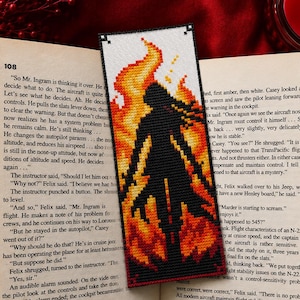 May include: A handmade bookmark featuring a black silhouette of a figure engulfed in flames, crafted with a cross-stitch design. The bookmark is placed in an open book with text visible on the pages, and a red candle is in the background.