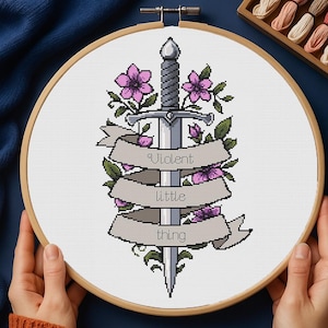 Könnte beinhalten: Kreuzstich-Stickerei in einem Holzrahmen mit einem Schwertdesign und dem Text "Violent little thing". Das Design umfasst lila Blumen und grüne Blätter auf weißem Hintergrund.