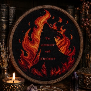 Può includere: Un ricamo a punto croce circolare in un telaio di legno che presenta una sagoma nera circondata da vivaci fiamme arancioni, rosse e gialle. Il testo "To whatever end" e "Fireheart" è ricamato con filo rosso al centro del disegno.