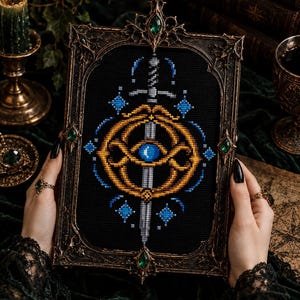 May include: A rectangular cross-stitch embroidery featuring a silver sword centred behind a golden circular emblem with a bright blue gemstone at its core. The design is set against a black fabric background and encased in an ornate dark metal frame.