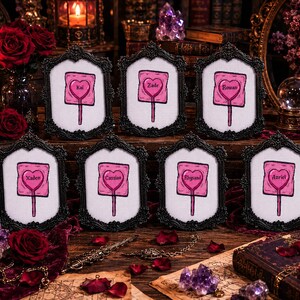 May include: Eight framed artworks with black ornate frames. Each frame holds a pink heart-shaped design on a white background, resembling a lollipop, with a name in the center. The names are Kai, Zade, Rowan, Xaden, Cassian, Rhysand, and Azriel.