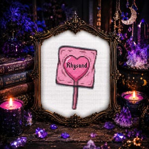 May include: A framed cross-stitch featuring a pink sign with a heart and the name "Rhysand." The sign is set against a white background within an ornate gold frame. The image is surrounded by books, candles, and crystals, creating a mystical aesthetic.