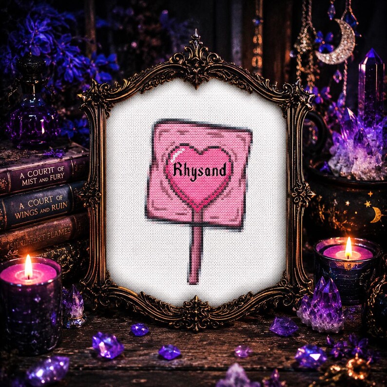 May include: A framed cross-stitch featuring a pink sign with a heart and the name "Rhysand." The sign is set against a white background within an ornate gold frame. The image is surrounded by books, candles, and crystals, creating a mystical aesthetic.