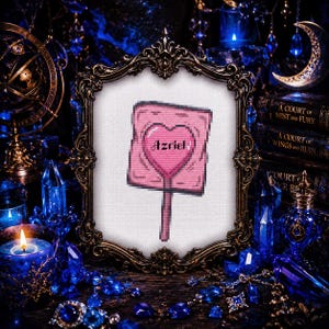 May include: A framed cross-stitch featuring a pink sign with a heart and the name "Azriel". The frame is ornate, with a dark, antique finish. The background includes blue crystals, candles, and books, creating a mystical aesthetic.