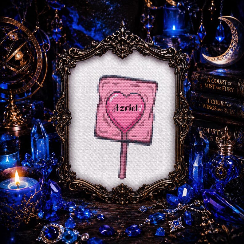 May include: A framed cross-stitch featuring a pink sign with a heart and the name "Azriel". The frame is ornate, with a dark, antique finish. The background includes blue crystals, candles, and books, creating a mystical aesthetic.