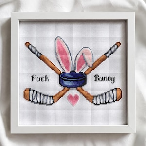 Könnte beinhalten: Ein gerahmtes Kreuzstich-Stickbild mit zwei gekreuzten Hockeyschlägern, einem dunkelblauen Puck und einem Paar rosa-weißer Hasenohren. Der Text "Puck" und "Bunny" ist beidseitig des Pucks gestickt, darunter befindet sich ein kleines rosa Herz.