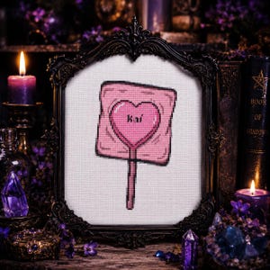 May include: A framed cross-stitch featuring a pink lollipop with a heart-shaped design and the name "Kai". The artwork is set against a white background and framed in a decorative black frame. The image has a gothic theme with candles and crystals.