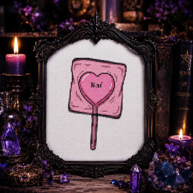 May include: A framed cross-stitch featuring a pink lollipop with a heart-shaped design and the name "Kai". The artwork is set against a white background and framed in a decorative black frame. The image has a gothic theme with candles and crystals.