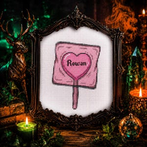 May include: Cross-stitch art piece featuring a pink sign with a heart-shaped center. The word "Rowan" is stitched inside the heart. The artwork is framed in an ornate, dark-colored frame, with candles and decorative elements in the background.