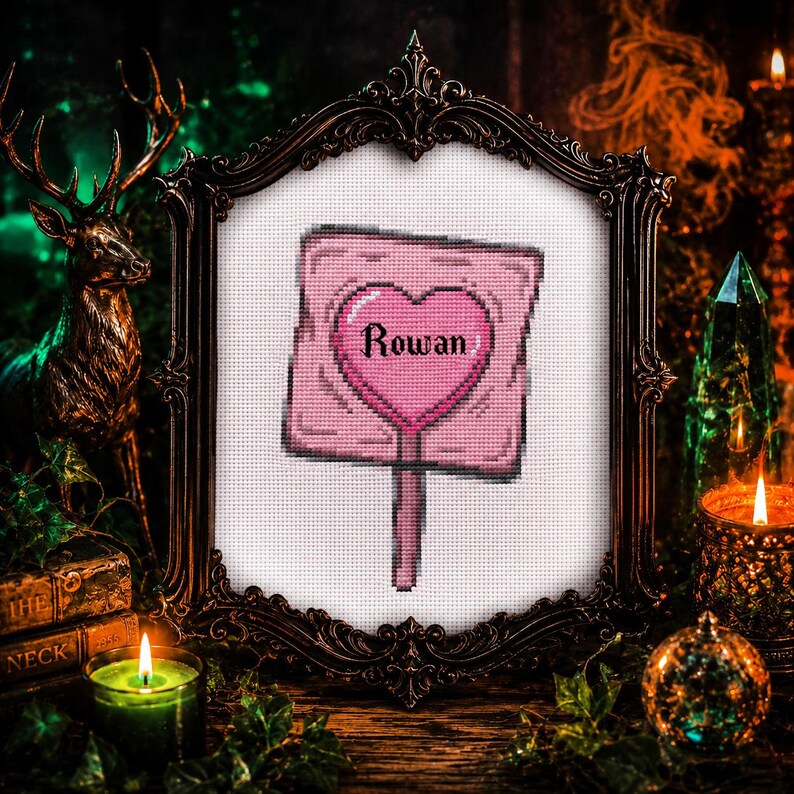 May include: Cross-stitch art piece featuring a pink sign with a heart-shaped center. The word "Rowan" is stitched inside the heart. The artwork is framed in an ornate, dark-colored frame, with candles and decorative elements in the background.