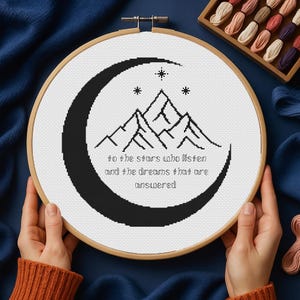 以下が含まれることがあります： 三日月と山が描かれた白黒のクロスステッチ刺繍のデザインで、"to the stars who listen and the dreams that are unanswered"というテキストが書かれています。