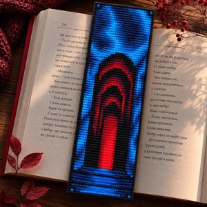 May include: A handmade bookmark with a pixelated design. The bookmark features a central red archway framed by a blue gradient, with a black border. The bookmark is placed on an open book with text.
