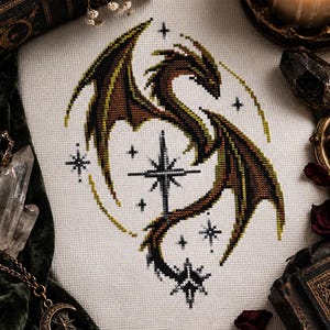 Puede incluir: Un bordado de punto de cruz sobre tela blanca que presenta un dragón con alas y cuerpo de color verde oscuro y marrón. El dragón está posicionado alrededor de una estrella central de cuatro puntas, con motivos de estrellas más pequeños en círculo.