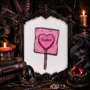 May include: A framed cross-stitch featuring a pink lollipop with a heart-shaped center and the name "Xaden". The frame is ornate, dark, and vintage-style. The background includes dragon figurines, candles, and books, creating a gothic aesthetic.