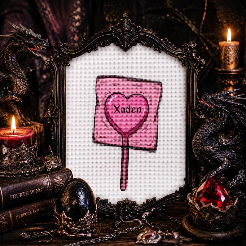 May include: A framed cross-stitch featuring a pink lollipop with a heart-shaped center and the name "Xaden". The frame is ornate, dark, and vintage-style. The background includes dragon figurines, candles, and books, creating a gothic aesthetic.