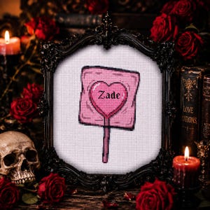 May include: A framed cross-stitch featuring a pink lollipop with a heart-shaped center. The word "Zade" is stitched in the heart. The frame is black and ornate, surrounded by red roses, candles, and a skull.