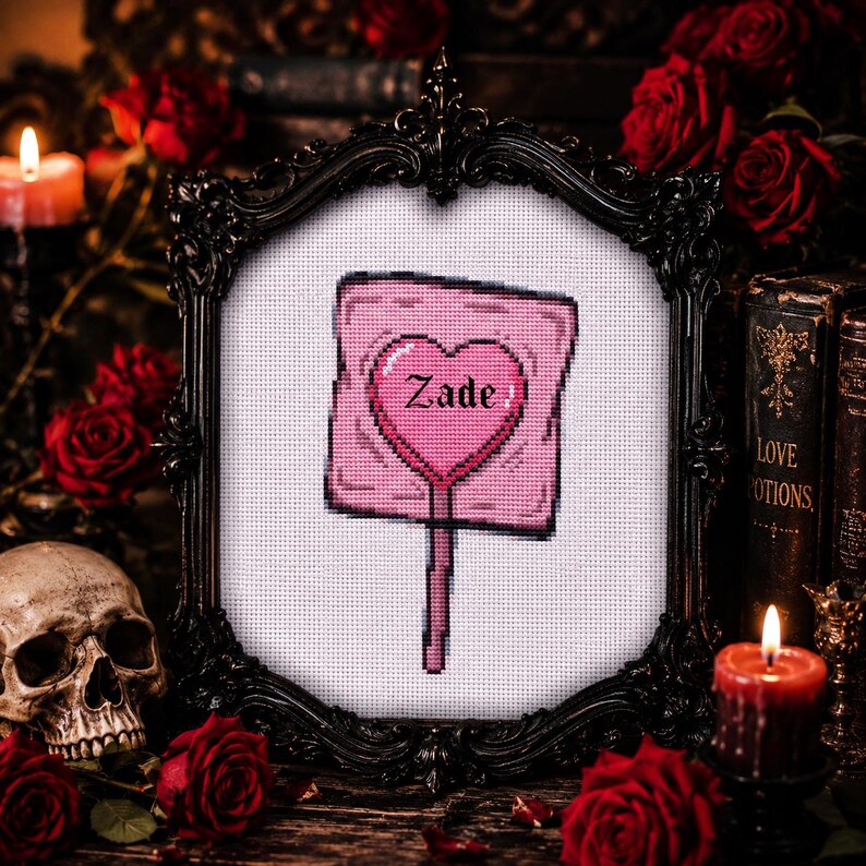 May include: A framed cross-stitch featuring a pink lollipop with a heart-shaped center. The word "Zade" is stitched in the heart. The frame is black and ornate, surrounded by red roses, candles, and a skull.