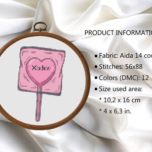 May include: A cross-stitch design of a pink lollipop with a heart-shaped center and the name "Xaden". The design is framed in a wooden embroidery hoop. The size of the design is 4 x 6.3 inches.