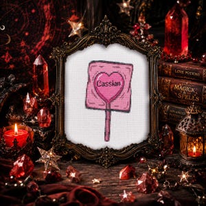 May include: A framed cross-stitch featuring a pink sign with a heart and the name "Cassian." The frame is ornate, dark, and sits on a wooden surface surrounded by crystals, candles, and books with titles like "Love Potions" and "Magick & Spells."