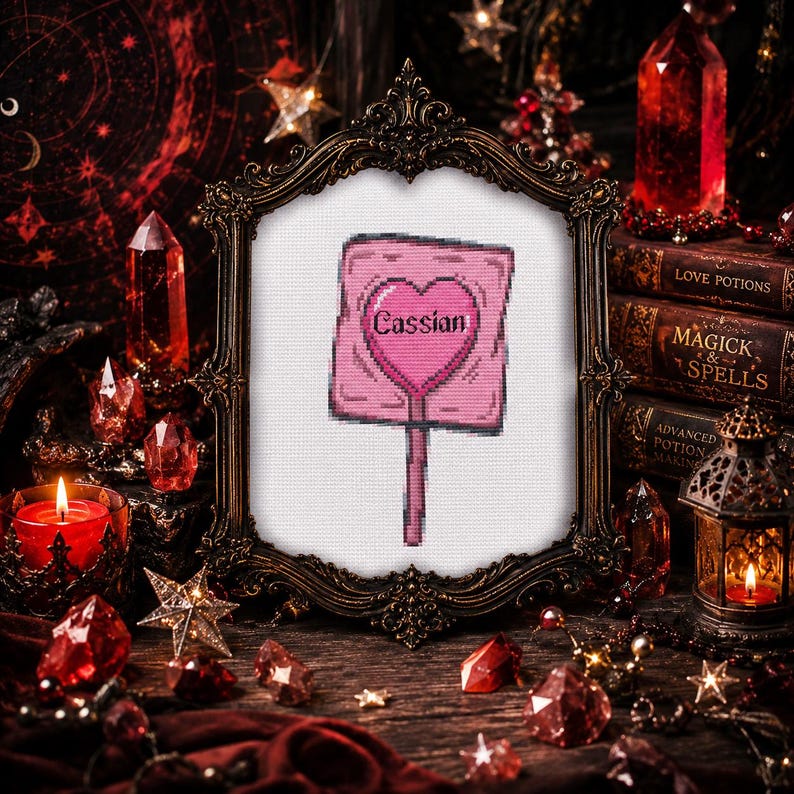 May include: A framed cross-stitch featuring a pink sign with a heart and the name "Cassian." The frame is ornate, dark, and sits on a wooden surface surrounded by crystals, candles, and books with titles like "Love Potions" and "Magick & Spells."