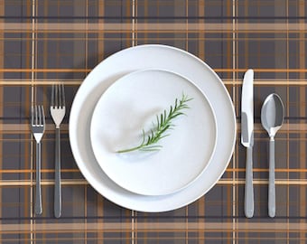 May include: A white dinner plate with a sprig of rosemary on top of a smaller white plate. The plates are set on a brown and orange plaid tablecloth with a silver fork and knife and a silver spoon.