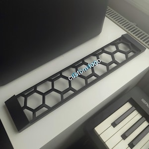 Op de afbeelding: Een zwart, rechthoekig decoratief paneel met een honingraatpatroon en de tekst "custom logo" in het midden. Het paneel ligt op een wit oppervlak, met een toetsenbord zichtbaar in de rechterbenedenhoek.