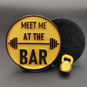 Peut inclure: Un badge de gym circulaire jaune et noir avec le texte "MEET ME AT THE BAR" et un graphique de barre. Un petit kettlebell doré est à côté du badge. Un objet circulaire noir est derrière le badge.