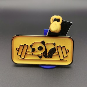 Op de afbeelding: Een gouden metalen pin met een zwart-witte pandabeer die een halterstang optilt. De panda ligt op zijn buik en heeft een schattige uitdrukking. De pin staat op een blauwe basis.