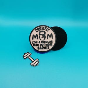 Puede incluir: Una pegatina redonda blanca y negra con el texto "Crossfit Mom Like a regular mom but more badass" y un icono de kettlebell negro. También hay un círculo negro y una mancuerna plateada en la imagen.