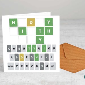 May include: A white greeting card with a Wordle-inspired design. The card features a grid with letter tiles in green, yellow, and gray, spelling out a word. An envelope in a brown color is next to the card.