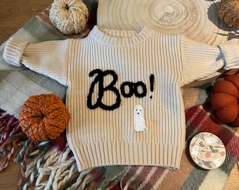 Puede incluir: Un suéter de punto beige con un "Boo!" bordado en negro en la parte delantera. El suéter está sobre una manta a cuadros con un pequeño letrero de madera con un diseño de fantasma y murciélago.